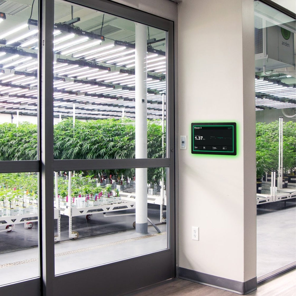 A smart indoor greenhouse environment with LED light arrays and plant trays visible through large glass doors, with a digital Growlink control panel mounted on a beige wall.
