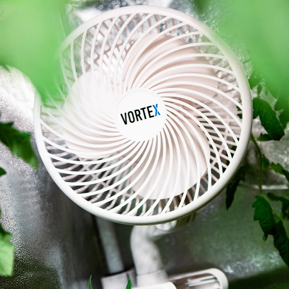 A close-up of a VORTEX fan with a white circular grille and white blades, shown in a real environment with green leaves and a mylar surface around it.