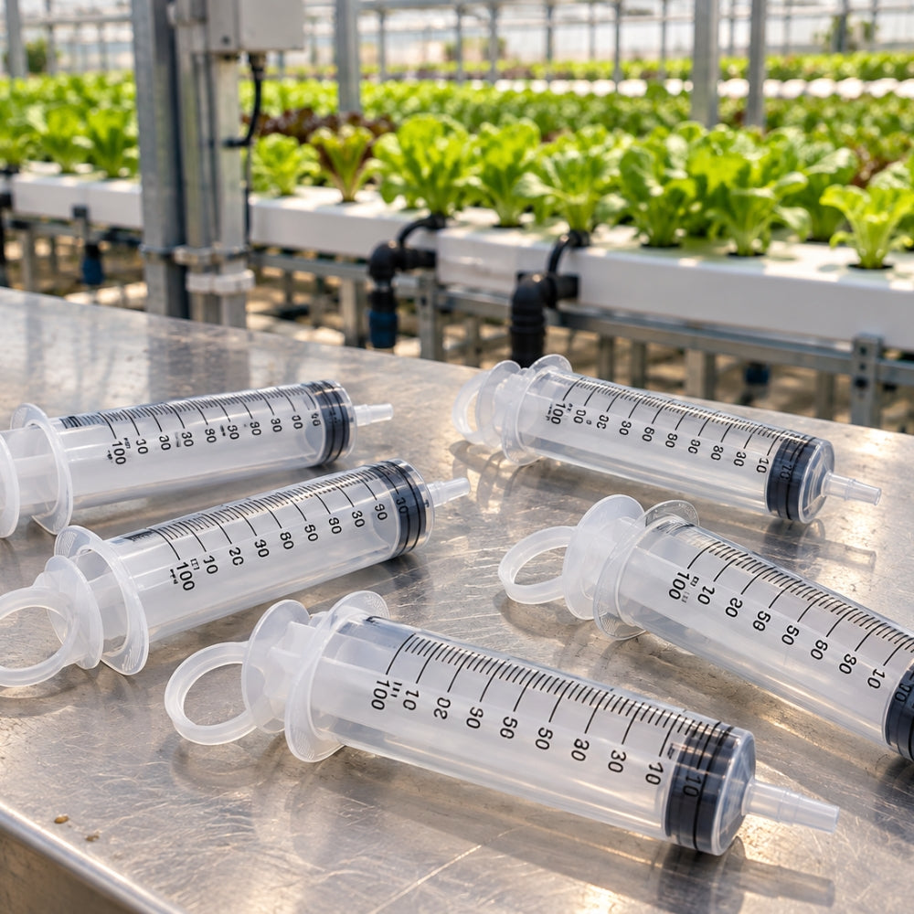 Five clear plastic syringes with measurement markings and black plungers arranged on a stainless steel table in a greenhouse with rows of green plants in the background.