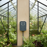 A dark gray Mission Control DigiStep Temperature Controller mounted on a light wood pillar inside a lush green greenhouse with glass panels.