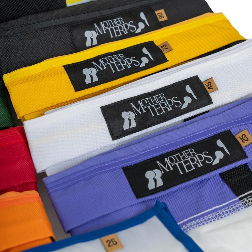 A close-up of several folded Mother Terps Bubble Bags stacked diagonally, showing the black woven Mother Terps logo patch on each bag. Visible bag colors include black, yellow, white, purple, red, orange, and blue, with small tan size tags.
