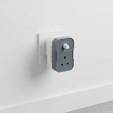 A Mission Controls BASiC-FAN fan controller plug-in, featuring a dark gray front faceplate with a white UK three-pin wall outlet and a white rotary speed control knob, set against a plain white background. shown plugged into a white wall socket.