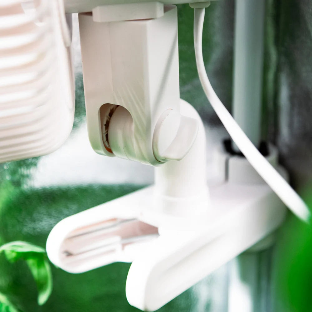 Close-up of a white plastic clip-on fan mount attached to a tent pole, with a white cord and blurred green background elements.