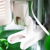 Close-up of a white plastic clip-on fan mount attached to a tent pole, with a white cord and blurred green background elements.